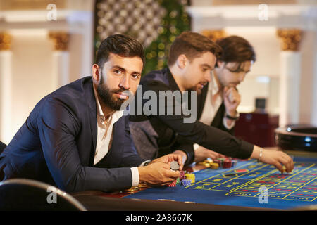 Un uomo in un vestito con un bicchiere di whiskey seduto al tavolo della roulette giocando a poker in un casinò. Foto Stock