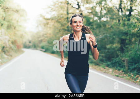 Giovani caucasici bruna atletica donna in sportswear corre una maratona su strada attraverso la foresta. Foto Stock