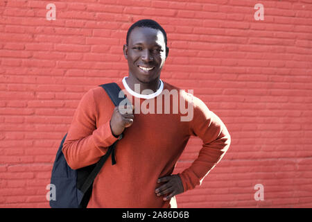 Ritratto di giovane africano un uomo Guarda la fotocamera e sorridente, portando allo zaino per la scuola. Copia spazio sulla Parete Rossa in background. Foto Stock