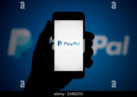 Un uomo guarda al suo iPhone che visualizza il logo di PayPal, con lo stesso logo in background (solo uso editoriale). Foto Stock