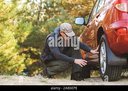 Uomo di sedersi vicino auto rossa controllare le ruote. Uomo anziano in tourist vestiti nella foresta. Foto Stock