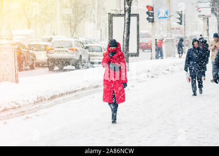 Una giovane donna in un cappotto rosso esegue il wrapping di se stessa e delle pelli dal vento. Tempesta di neve in un ambiente urbano. Abstract inverno sfondo meteo Foto Stock