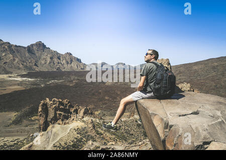 Uomo sportivo con zaino e gode di una bellissima giornata dopo la scalata da solo. Ragazzo caucasico guardando lontano seduto sulla scogliera di montagna che si affaccia sul panorama Foto Stock