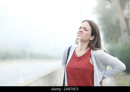 La donna soffre del mal di schiena in piedi un nebbioso giorno freddo in un parco Foto Stock