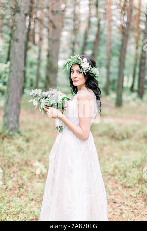 Bella Sposa con bouquet di fiori in abito bianco si erge su sfondo di foresta. Stile rustico Foto Stock