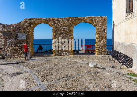 Nei pressi del molo a Cefalú, Sicilia, Italia, un uomo stand vende la sua mercanzia di fronte ad un doppio arco che conduce al mare. Foto Stock