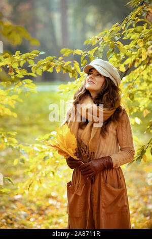 Ciao autunno. donna moderna in un maglione, gonna, cappello, guanti e sciarpa con foglie di giallo alla ricerca fino a copiare lo spazio all'aperto nel parco di autunno tra le Foto Stock