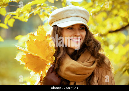 Ciao autunno. sorridente giovane donna in maglia, cappello e sciarpa con foglie di giallo all'aperto nel parco di autunno tra le fronde. Foto Stock