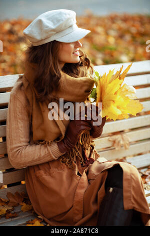 Ciao autunno. sorridente giovane donna in maglia, gonna, cappello, guanti e sciarpa con foglie di giallo guardando in lontananza mentre è seduto su una panchina superarvi Foto Stock