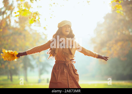 Ciao autunno. allegro moderno 40 anno vecchia donna in maglia, gonna, cappello, guanti e sciarpa con foglie di giallo all'aperto nel parco d'autunno. Foto Stock