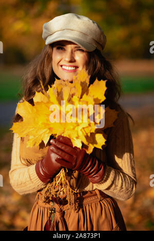 Ciao autunno. Ritratto di felice alla moda di 40 anno vecchia donna in maglia, gonna, cappello, guanti e sciarpa con foglie di giallo guardando in lontananza per esterno Foto Stock