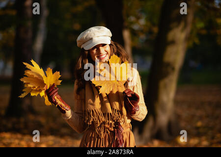 Ciao autunno. Ritratto di sorridere moderno medio evo woman in maglione, gonna, cappello, guanti e sciarpa con foglie di giallo al di fuori del parco d'autunno. Foto Stock
