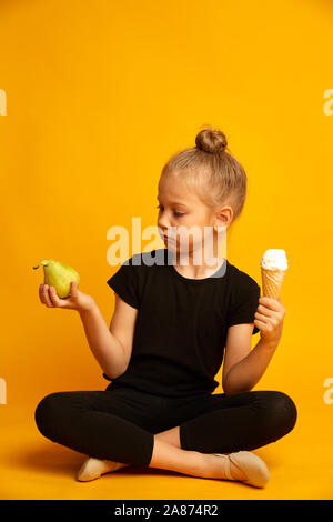 Incantevole piccola ragazza bionda scegliendo tra la pera e il dolce di gelati su sfondo giallo Foto Stock