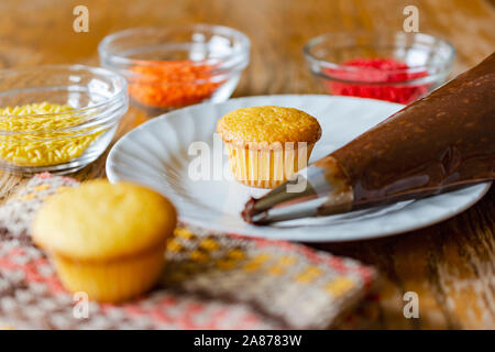 Mini cupcake sulla piastra bianca con glassa di cioccolato in tasca da pasticciere sfocato in primo piano. Giallo, arancione, rosso e spruzza in ciotole in vetro sfocato nella Foto Stock