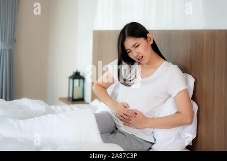 Donna asiatica infelice giacente sul letto cercando di malessere, soffre di mal di stomaco in camera da letto, stomachache a causa delle mestruazioni e mangiare rovinato fo Foto Stock