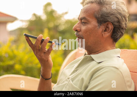 Senior uomo utilizzando assistente virtuale nel telefono - Indiana uomo anziano utilizzando il comando vocale sul cellulare - vecchio uomo che parla sullo smartphone all'aperto. Foto Stock