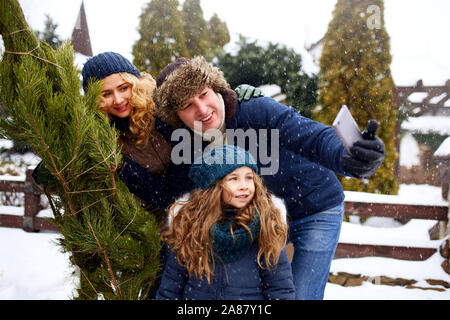 Bellissimo mamma, papà e figlia facendo selfie con taglio fresco albero di natale con lo smartphone vicino a casa. Giovane famiglia Buon Natale e Felice nuovo Foto Stock