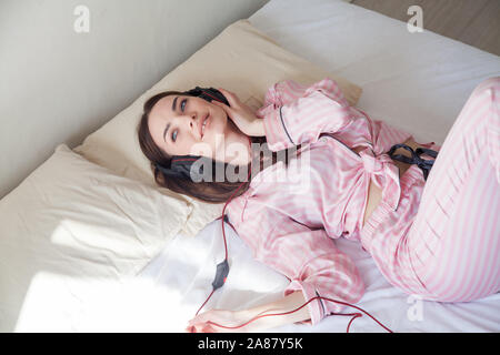 Donna rosa in pigiama ascolta la musica con le cuffie sul letto Foto Stock