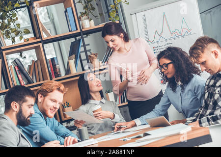 Startupers lavorando insieme a Office seduta che mostra una donna incinta collega piano di progetto allegro Foto Stock