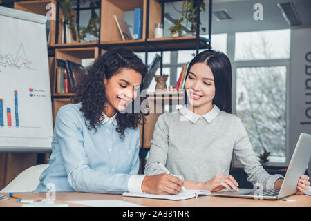 Startupers femmina lavorando insieme a Office seduta la navigazione portatile prendendo appunti allegro Foto Stock