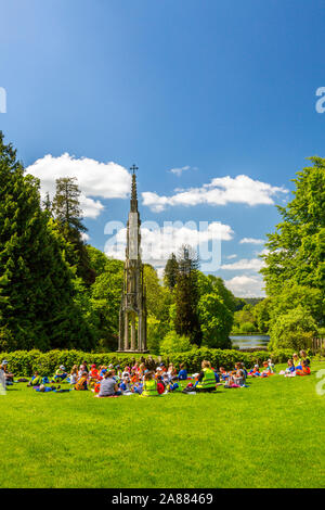 Un gruppo di bambini della scuola di mangiare il pranzo di fronte alla ex Bristol alta croce di giardini Stourhead, Wiltshire, Inghilterra, Regno Unito Foto Stock