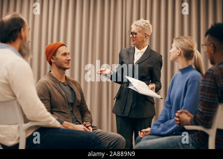 Imprenditrice maturo con corti capelli biondi e in tuta in piedi e a parlare con le persone che ha premuto il seminario Foto Stock