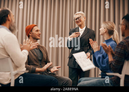 Gruppo di persone seduti su sedie e plaudendo alla donna matura per la sua performance di successo Foto Stock