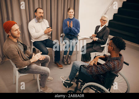 Gruppo di persone seduti su sedie in cerchio e battendo le mani essi saluto il loro nuovo membro del gruppo Foto Stock