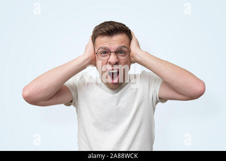 Infastiditi giovane uomo in bicchieri urlando e coprendo i suoi orecchi perché egli non può sopportare il rumore. Viso negativo emozione umana Foto Stock