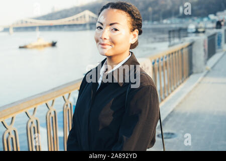 Una giovane e bella ragazza di etnia africana con la vitiligine in piedi sulla calda primavera città strada vestito mantello nero close up ritratto di donna con s Foto Stock