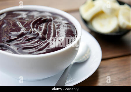 Ciotola fresca di viola acai amazzonico con fettine di banana in un bianco pulito ciotola sul tavolo di legno in Brasile Foto Stock