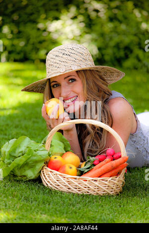 Blonde junge Frau liegt auf einer Wiese im Park und geniesst Obst und Gemüse, Picknick, Strohhut, Foto Stock