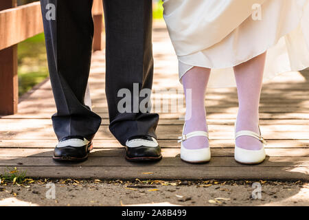 I piedi di una sposa e lo sposo in piedi insieme su un ponte il giorno delle nozze. La sposa si solleva il suo abito di rivelare i suoi collant e scarpe di bianco. Foto Stock