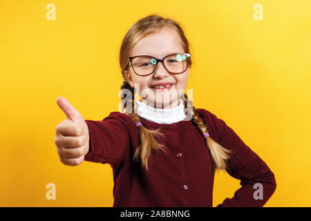 Bambino studente con gli occhiali mostra il pollice verso l'alto. Bambina con pig-tail in un maglione di borgogna su sfondo giallo. Foto Stock