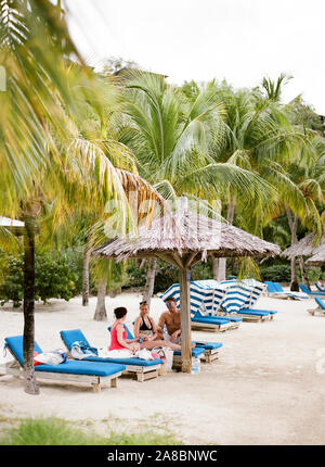 Amici adulti seduti sotto palm frond ombrelloni sulla spiaggia al Bitter End Yacht Club di Virgin Gorda Isola, Isole Vergini Britanniche Foto Stock