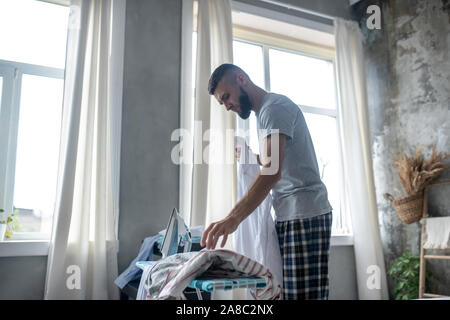 Uomo Barbuto in piedi vicino alla finestra e servizio stireria Foto Stock