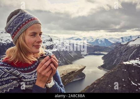 Giovane donna in norvegese maglione bevande di tè, gode di vista su Trolltunga, Norvegia Foto Stock