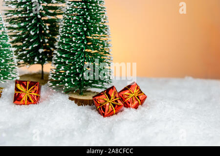 Close-up di rosso regali di Natale sulla neve sotto piccolo giocattolo di abeti. Illuminata luminosamente regali di natale nella foresta di giocattolo. Merry Christmas greeting card e ho Foto Stock