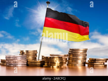 Bandiera tedesca con pile di monete metalliche in euro nella parte anteriore del cielo blu Foto Stock
