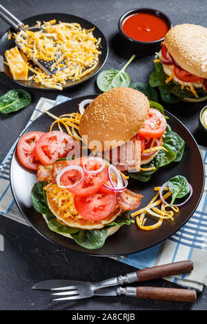 Hamburger di pollo su un panino con marmo shredded formaggio fritto croccante pancetta, le fette di pomodoro, foglie di spinaci e cipolla rossa su una piastra nera, close-up, vert Foto Stock