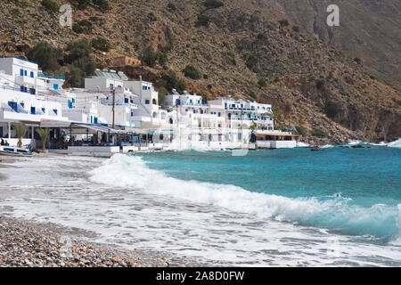 Loutro, la Canea, Creta, Grecia. Onde che si infrangono sulla spiaggia di pietra, taverne sul lungomare sullo sfondo. Foto Stock