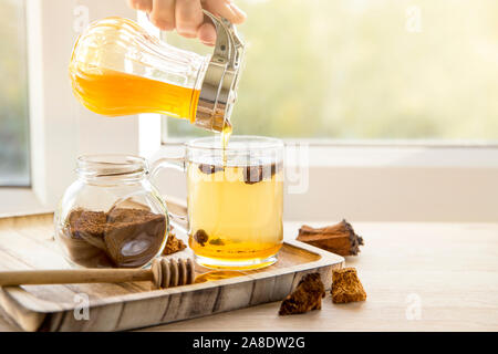 Puro sano selvaggio chaga fungo betulla, Inonotus obliquus pezzi in vetro di tè sul vassoio in legno sul davanzale, soleggiata luce posteriore. Foto Stock