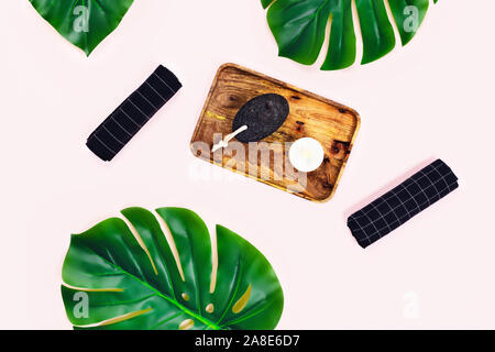 Sfondo rosa con vassoio in legno nero e asciugamani, tazza di caffè espresso e pietra pomice circondato da foglie tropicali. Appartamento laico, vista dall'alto. Concetto di Spa. Foto Stock