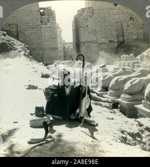 'Un Egiziano serpente incantatore pile il suo commercio accanto al viale di Sfingi, Tempio di Karnak, Tebe, Egitto", c1930s. Tempio di Karnak complesso nei pressi di Luxor in Egitto ha iniziato durante il regno di Senusret I nel Regno di Mezzo e continuato nel periodo tolemaico. Il viale di Sfingi collegato i Templi di Luxor e Karnak e fu usato per le processioni cerimoniali. Da "Giro del mondo". [Keystone View Company, Meadville, Pa., New York, Chicago, Londra] Foto Stock