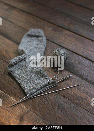 Mano calza a maglia con aghi e filo grigio sulla sfera di legno scuro dello sfondo. Concetto di fatto a mano e hugge slow life. Foto Stock