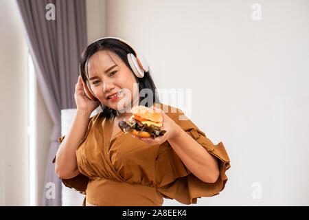 Fame sovrappeso donna utilizzando le cuffie è sorridente e tenendo un hamburger e seduto in soggiorno, la sua molto felice e gode di mangiare fast food Foto Stock