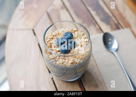 Mirtillo parfait di yogurt con muesli, avena e chia semi in un vetro bianco su un tavolo di legno. Colazione sana. close up Foto Stock