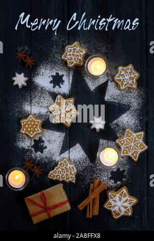 Merry Christmas testo biglietto di auguri con gingerbread cookies, spezie e candele su una stella da zucchero a velo sul blu scuro dello sfondo di legno, alto angolo Foto Stock