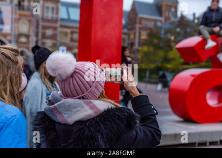 Ragazza scatta una foto del fidanzato sul mio segno di Amsterdam di Amsterdam, di fronte al Rijksmuseum con il suo cellulare in effetto droste indossando un n Foto Stock