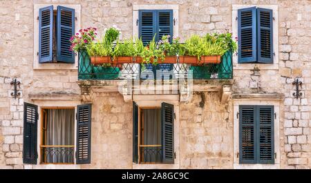 Vista sulla strada di una bella casa in pietra con facciata dipinta di persiane alle finestre e un piccolo balcone con una cancellata in ferro battuto e di piante in vaso. Montenegro. Foto Stock